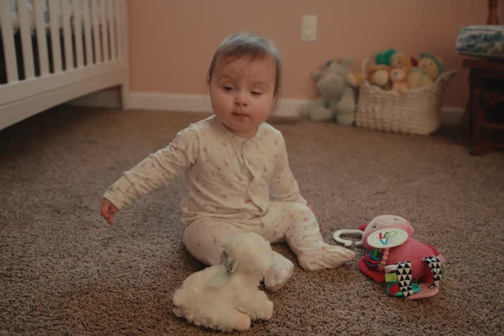 A baby sits on a carpeted floor surrounded by toys in a cozy room