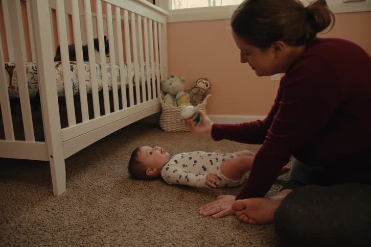 A mother sits on a carpeted floor feeding a baby lying on its back with a bottle next to a white crib in a softly lit room