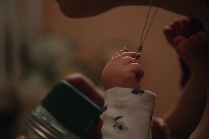 A persons profile likely a mother as a babys hand playfully interacts with a thin chain hanging from the persons neck the scene has a warm intimate feel with soft lighting and a blurred background