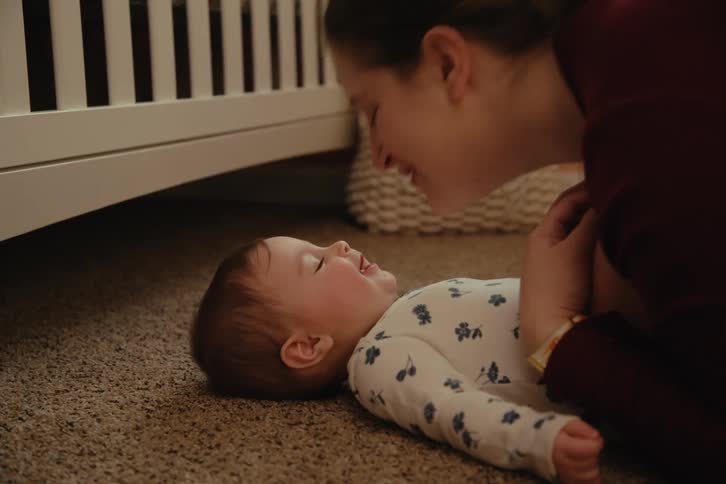 Mother playing and kissing her baby girl who is lying on the carpet in a room