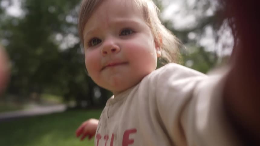 A baby reaches toward the camera with wide eyes and a soft expression set in a grassy backyard or park