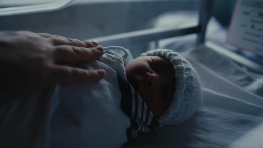 A closeup of a newborn baby swaddled in a blanket and wearing a light blue knitted cap lying in what appears to be a hospital bassinet a hand gently touches or caresses the babys head conveying a sense of tenderness and care