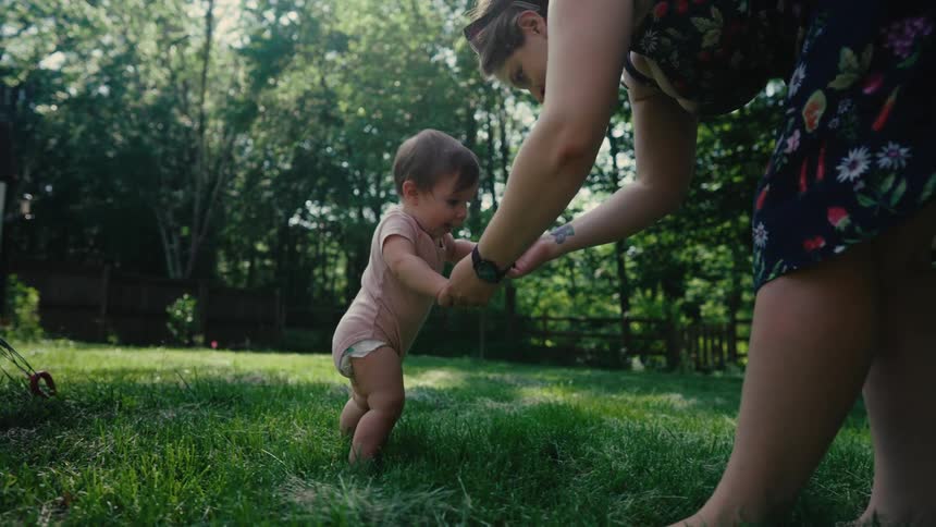 A mother helps her baby take steps on the grass in a sunlit backyard