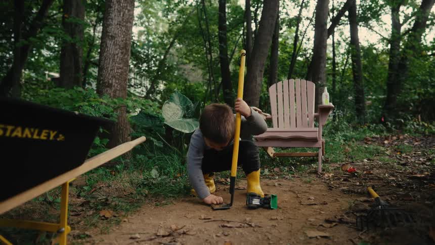 Young boy in yellow boots playing with tools in a forest clearing pretending to dig