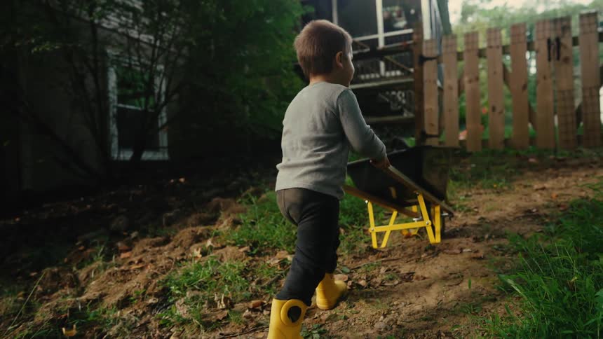 Little boy in boots playing with a construction wheelbarrow in a natural area