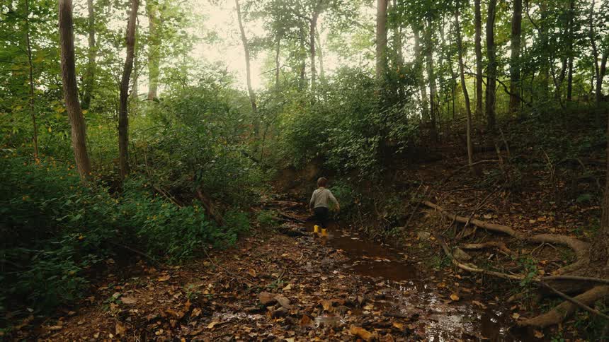 Little boy in boots walking along a trail beside a small river