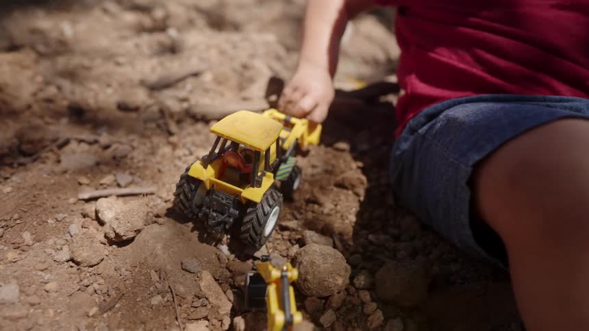 Male toddler plays with toy vehicles while sitting on a soil heap outdoors during daytime