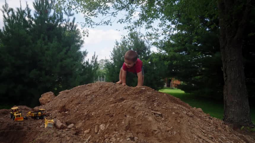 The boy climbs on the dirt near his toy trucks