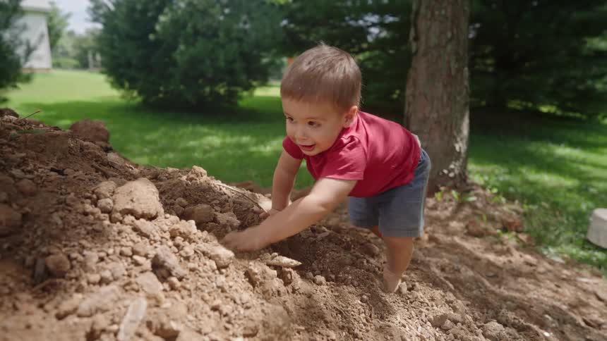 A toddler wearing jean shorts and a red shirt crawls on a sand hill on a lawn at midday