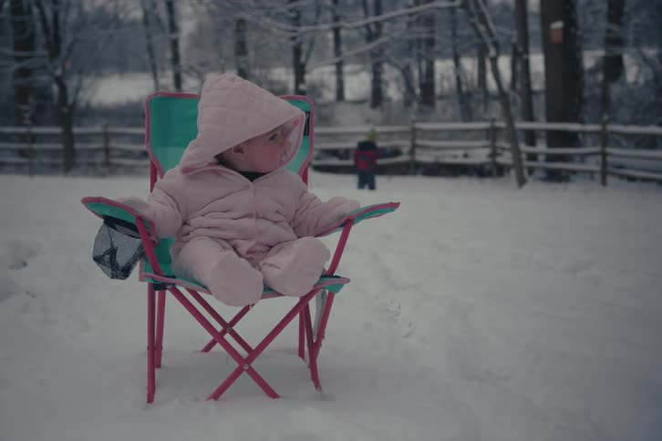 Little girl sitting on a chair in a snowy environment and in the background another child playing