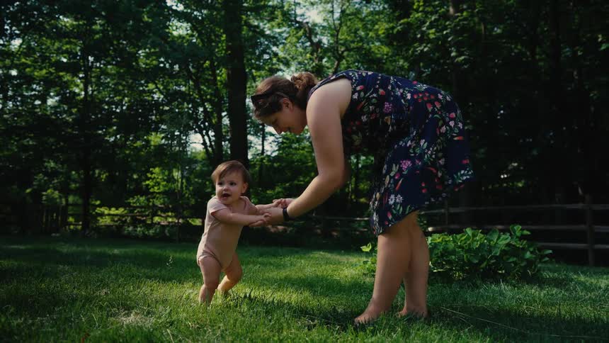 Mother guides baby as he takes early steps on a lawn outdoors during daytime
