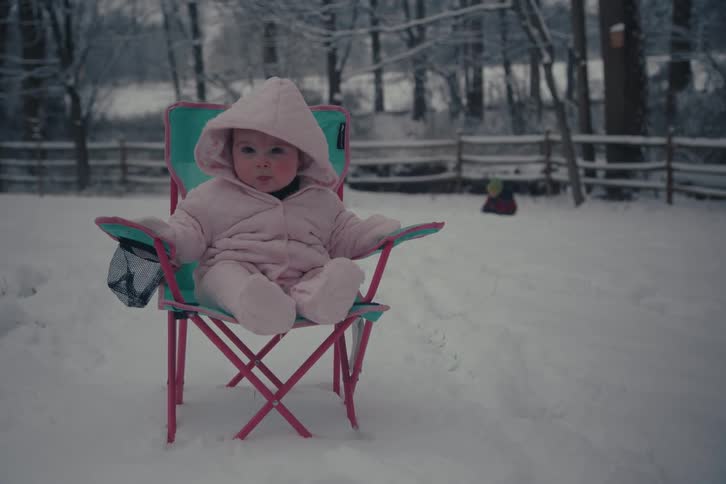 A bundledup baby in a pink snowsuit sits in a small chair on a snowy lawn with a wintry forest and another child playing in the background