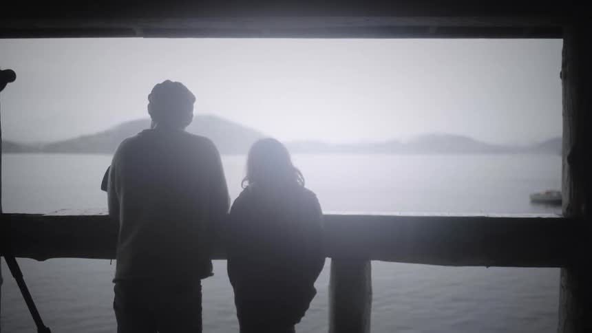Two people stand under a shelter looking out over a calm lake and distant mountains