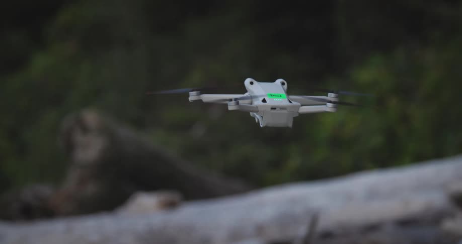 A small white drone with green lights is flying over a pile of large fallen tree trunks in a forested area