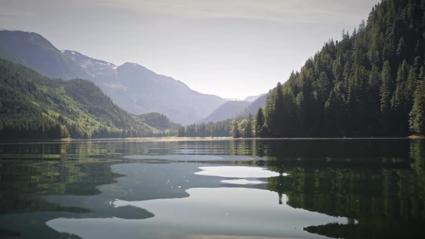 A vast body of calm water reflects the surrounding forested mountains and a bright hazy sky