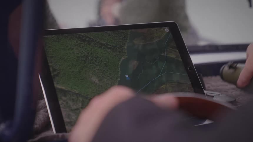 A person in the drivers seat of a car holds the steering wheel with one hand with a gps screen in front and picks up a monocular device at midday