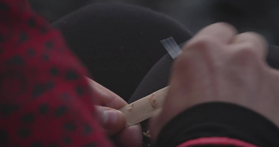 A closeup of hands meticulously scraping a wooden stick with a sharp edge generating small shavings this action suggests a detailed craft or woodworking activity is taking place
