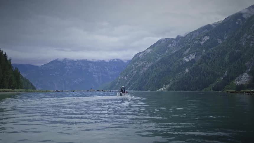 A small motorboat travels across the water moving away from the camera toward steep forested mountains under a cloudy sky