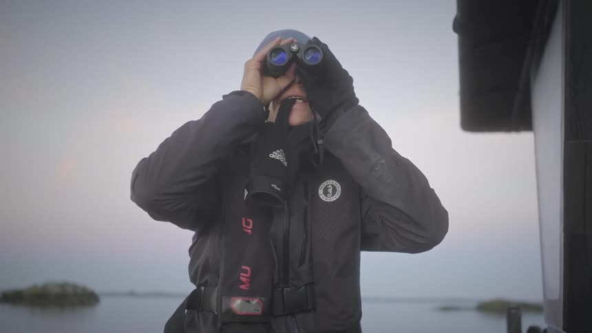 A person in a dark jacket and blue beanie looks through black binoculars at a calm body of water beneath an overcast sky