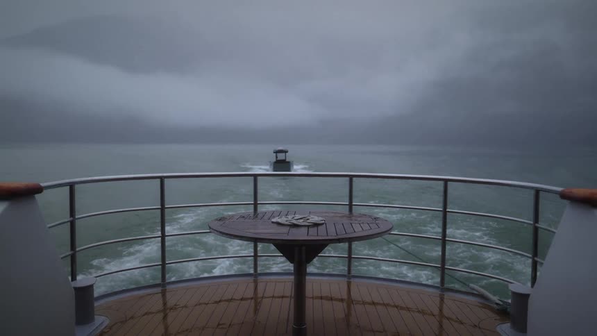 Viewed from the stern of a large boat a smaller vessel is towed across the water through dense fog with misty mountains visible in the distance