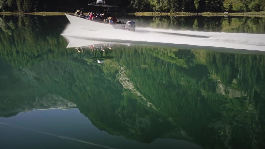 A sleek white motorboat cuts across a tranquil mirrorlike lake leaving a clear wake behind it the surrounding majestic treecovered mountains are perfectly reflected on the waters surface creating a stunning symmetrical landscape