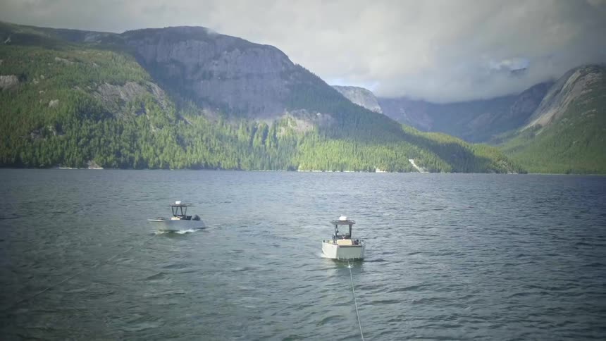 Two large boats leave wakes while being towed across the water away from the forested mountainous shore on a foggy day