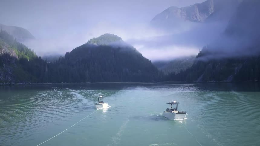 Two small boats glide across a misty lake surrounded by forested mountains