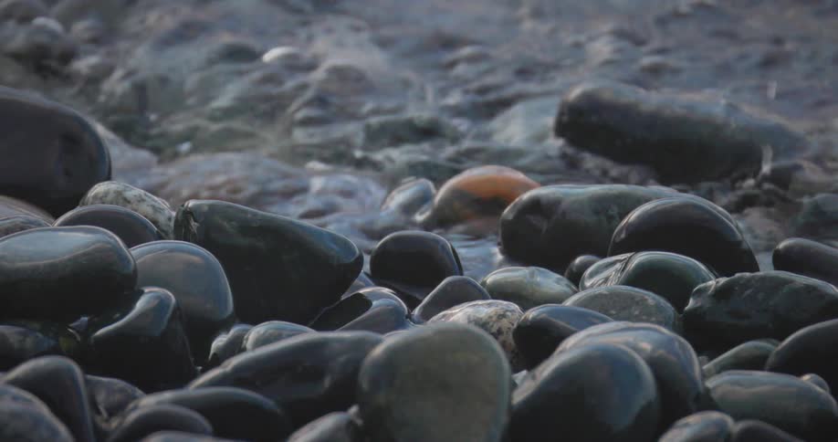A closeup shot captures gentle waves washing over smooth dark rocks on a shoreline