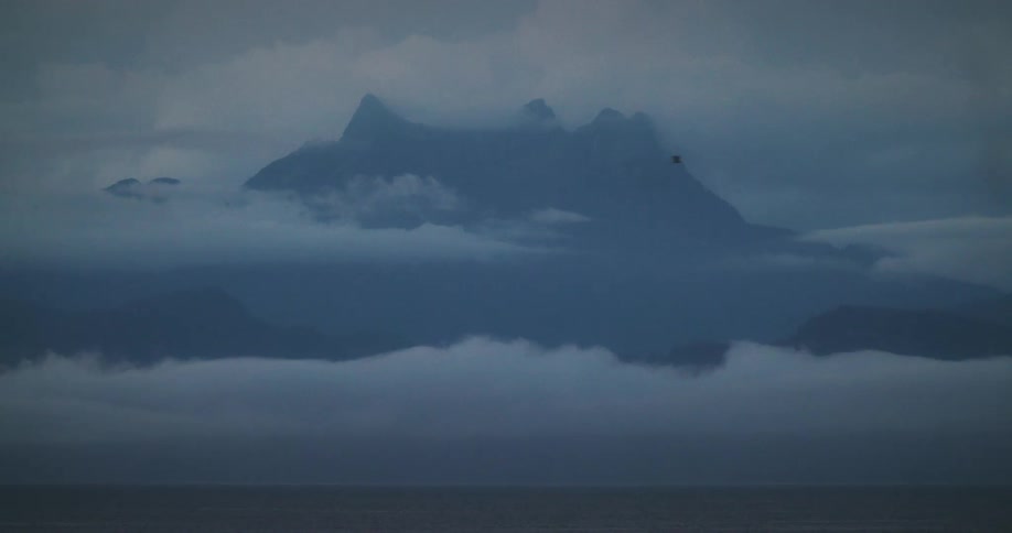 A jagged mountain peak rises through layers of mist and clouds over a calm body of water creating a moody and atmospheric scene