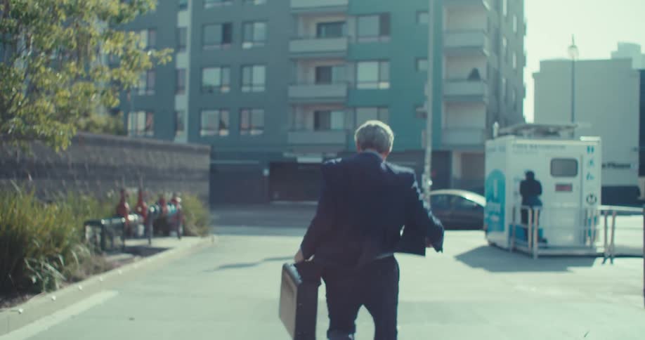 A man in a suit walks away down a city street holding a briefcase and raising his hands in happiness