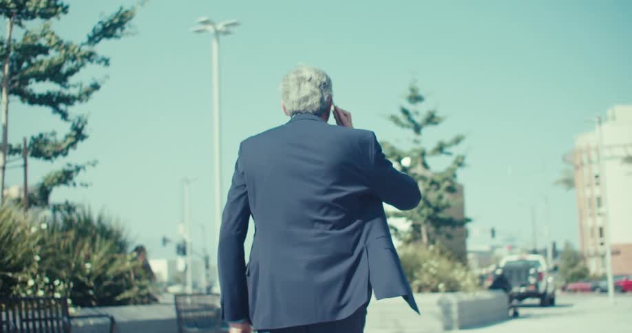 An older man in a suit walks through a sunny urban plaza while talking on the phone and carrying a briefcase
