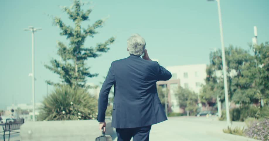 An older man in a suit walks outdoors talking on his phone under a clear blue sky