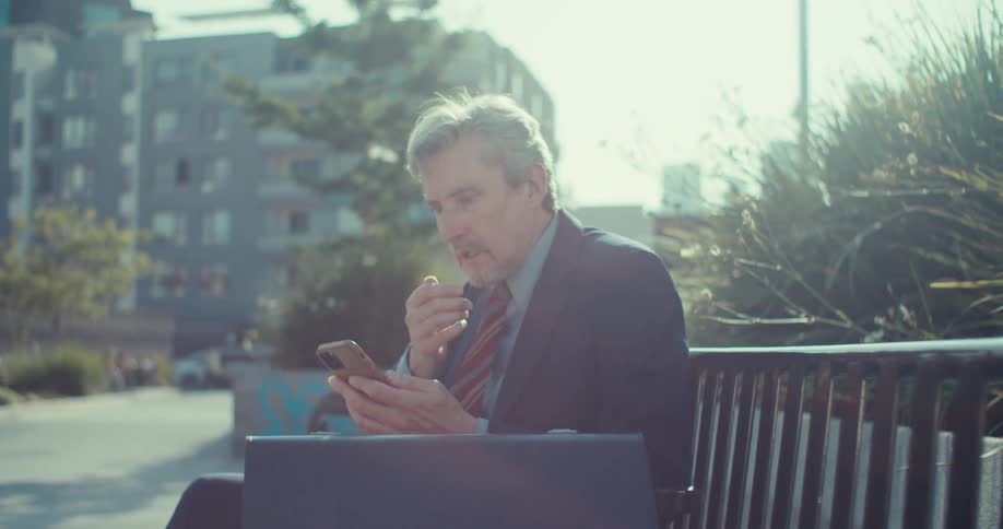 A man sits on a bench in the park scrolling through his phone then suddenly looks surprised and sad at what hes seeing at midday