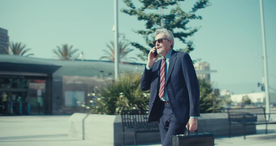 A welldressed man carrying a briefcase and wearing sunglasses walks down a sunny city street while talking on his phone he appears to be a businessman commuting or on a workrelated call