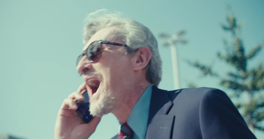 An older man in a suit and sunglasses stands outdoors under a clear blue sky looking downward with a serious expression