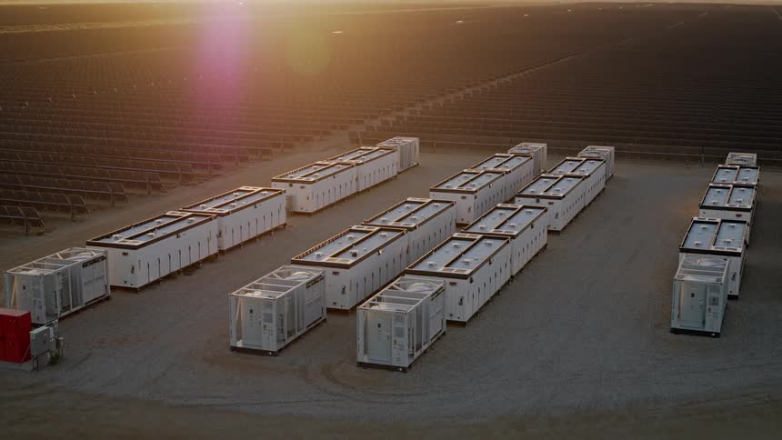 A large aerial array of white battery units stretch across a flat arid landscape of solar panels