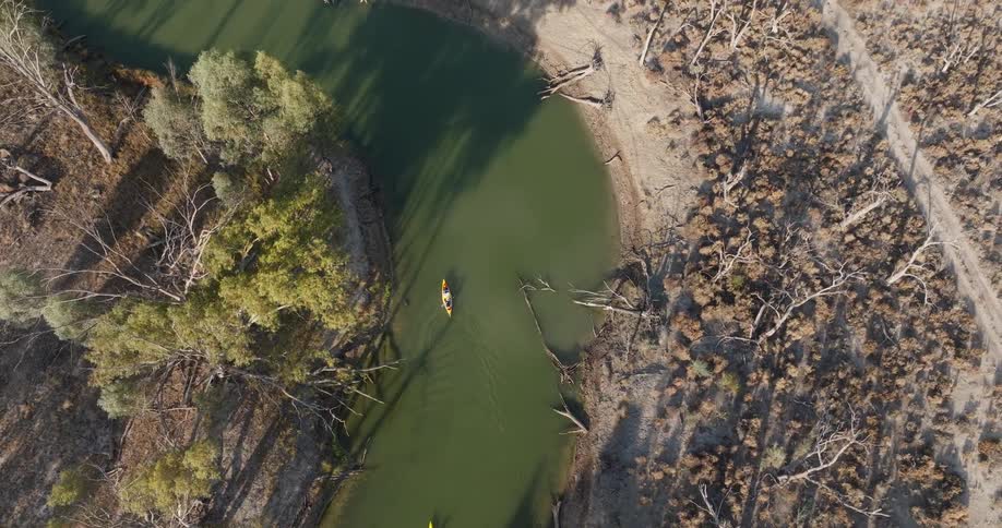 Two kayakers paddle down a winding green river through a dry wooded landscape