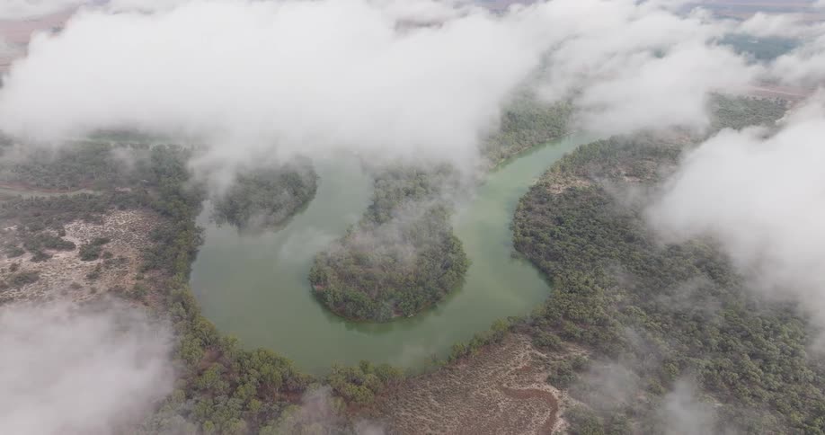 An aerial shot looks down through white clouds at a winding river that curves around a small forested island