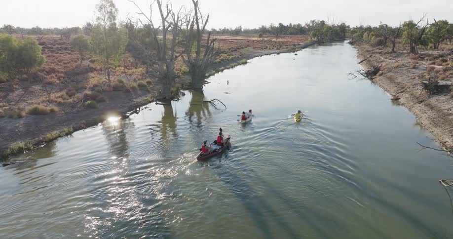 Three kayaks with people paddling along a calm river surrounded by dry vegetation and trees
