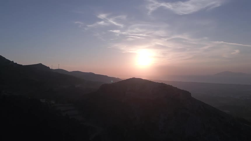The sun sets behind a jagged mountain ridge casting a golden glow across a hazy sky and silhouetted peaks