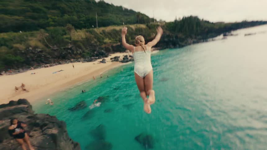 Girl jumps from a cliff into the ocean near a beach on an overcast summer day