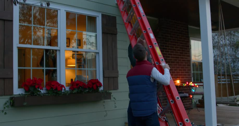 A man helps steady a tall ladder next to a house decorated for the holidays