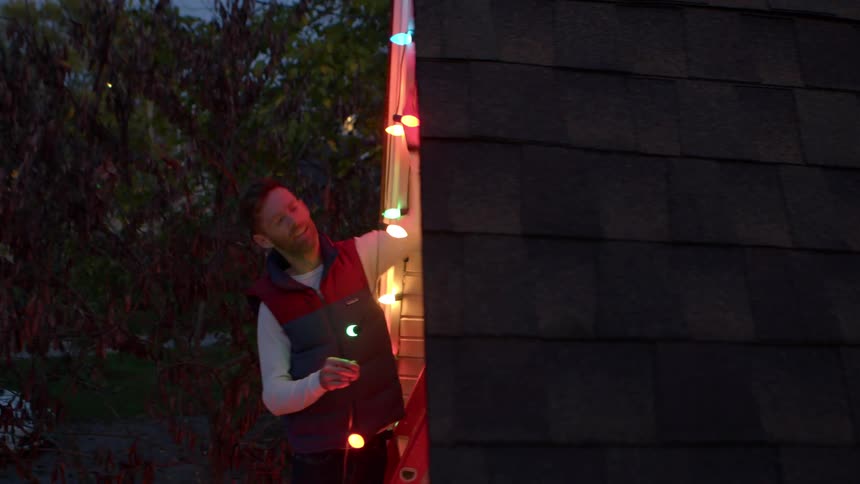 Man standing on a ladder puts up a string of colored lights on his home exterior