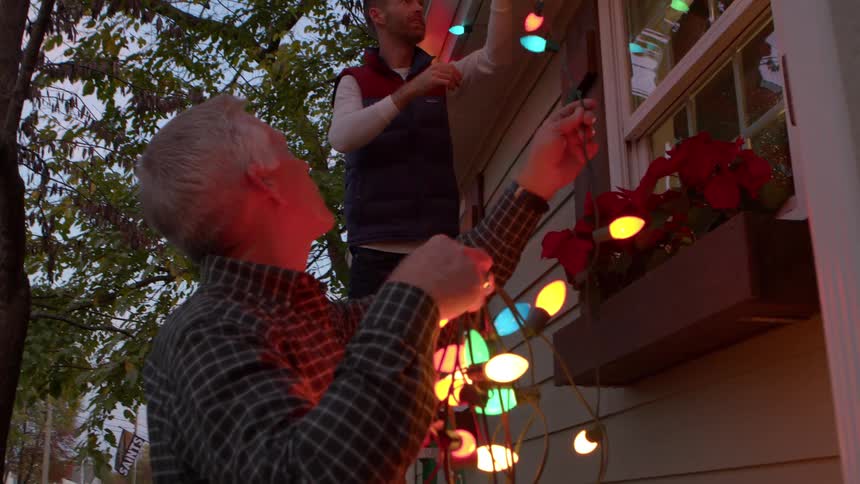 Adult son hanging up christmas lights on house as dad holds up the ends