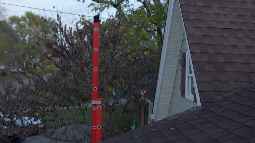 A long red ladder extends towards the top of a house with a brown shingled roof and light colored siding with bare tree branches visible in the background
