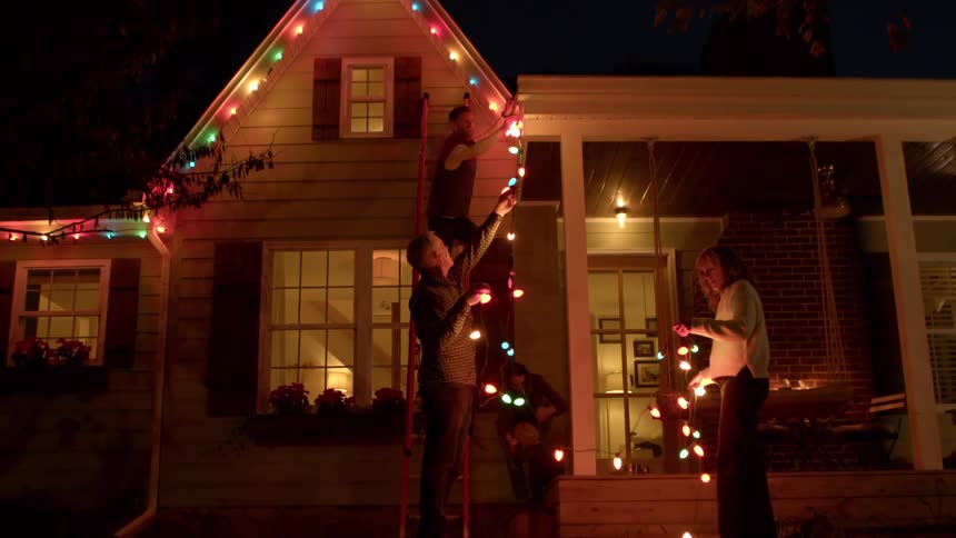 Adult son hanging up christmas lights on parents house as they hold the ends