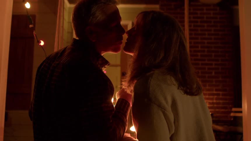 Two people kiss while hanging string lights in a dimly lit outdoor space with warmtoned patio furniture in front of a house
