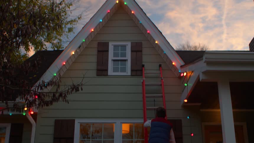 Man climbing a ladder towards a window with the house decorated with christmas lights