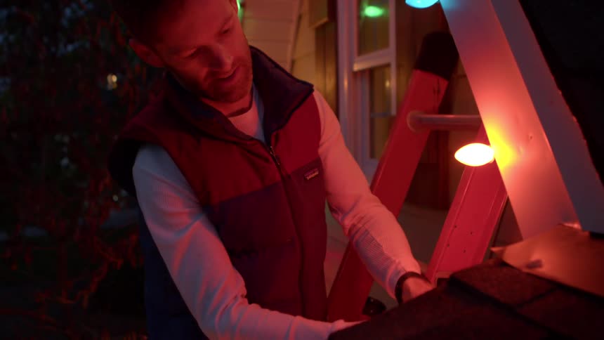 A man with a beard wearing a vest over a longsleeved shirt standing outdoors in the evening he is actively involved in setting up or adjusting colorful string lights on a house with a ladder visible nearby
