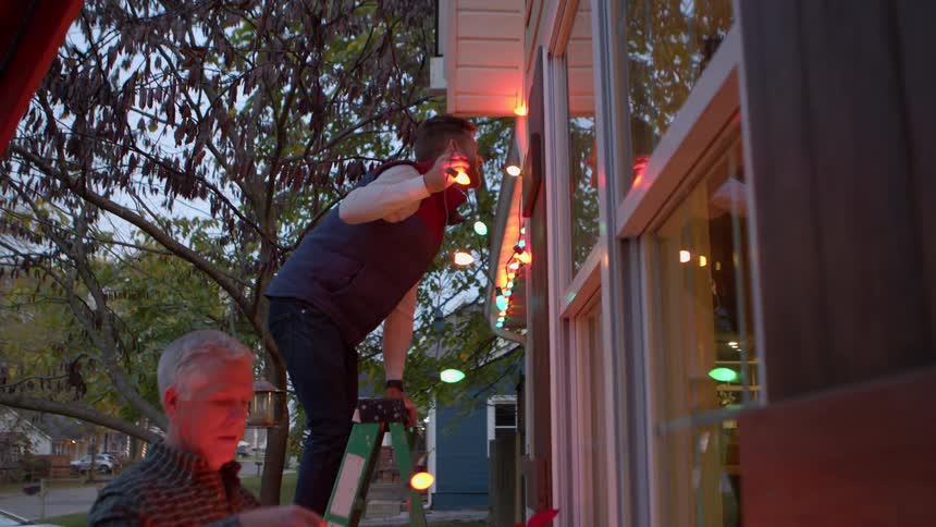 Adult son climbing ladder to hanging up christmas lights on house as dad untangles the ends